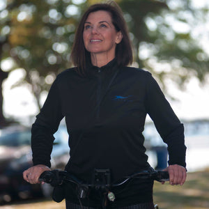 Smiling, brown-haired woman on bicycle in a park wearing Herron Logo Collection pullover