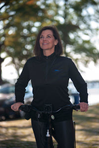 Smiling, brown-haired woman on bicycle in a park wearing Herron Logo Collection pullover