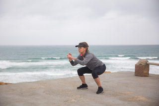 Mature woman in chair yoga pose on a concrete pier by the ocean wearing Herron Apparel leggings and eco-friendly jacket.