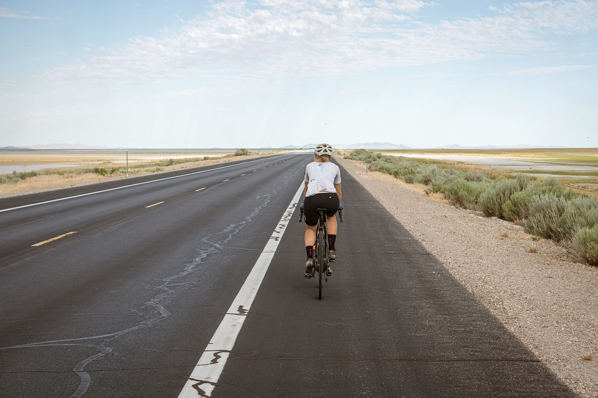 Mature woman from behind riding a bike on a flat road through the desert wearing Herron Apparel cycling shorts and jersey.